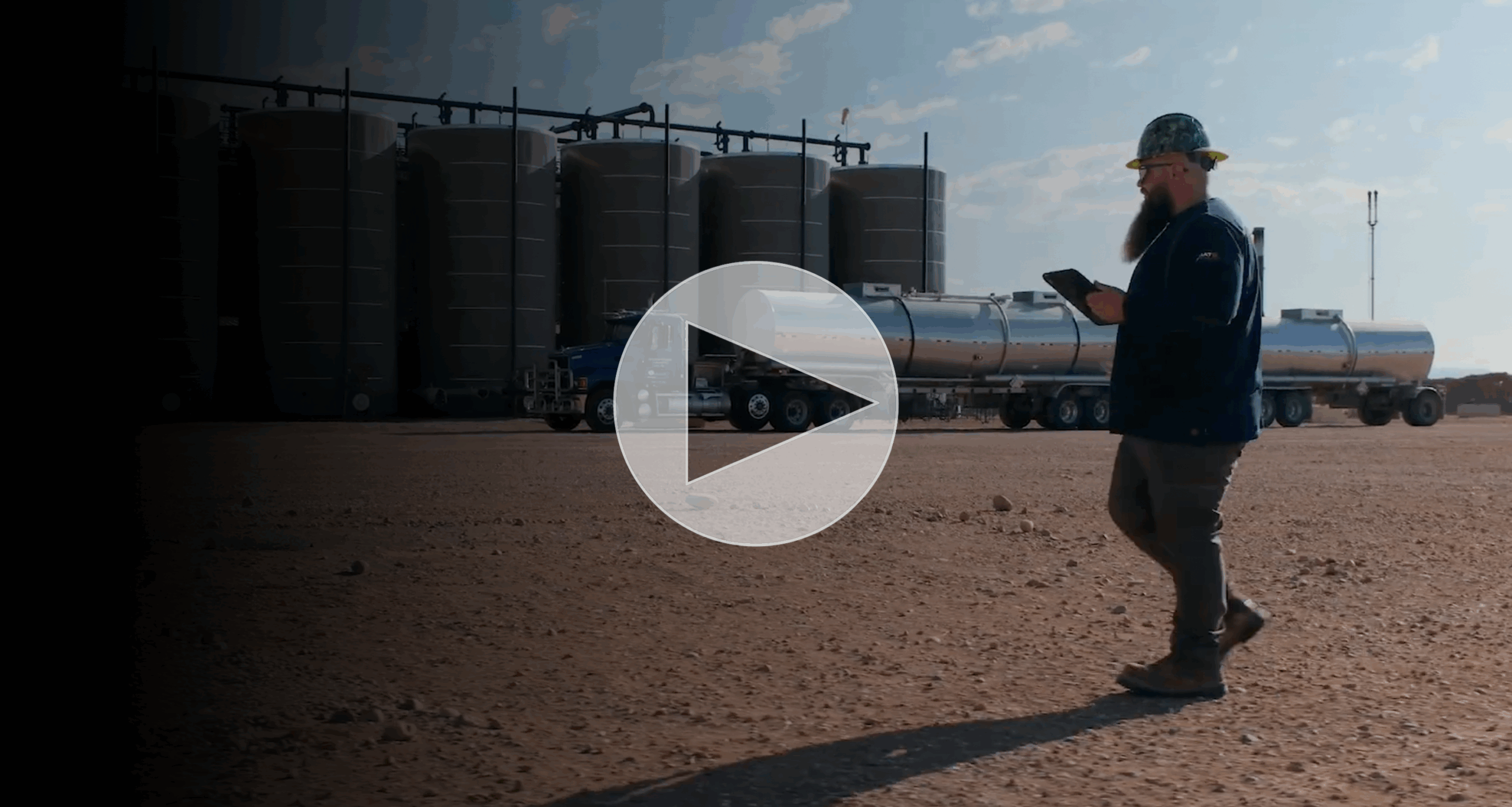 Thumbnail for a video showing a man with a beard and a hard hat walking and looking at a tablet in front of large industrial tanks and a tanker truck.