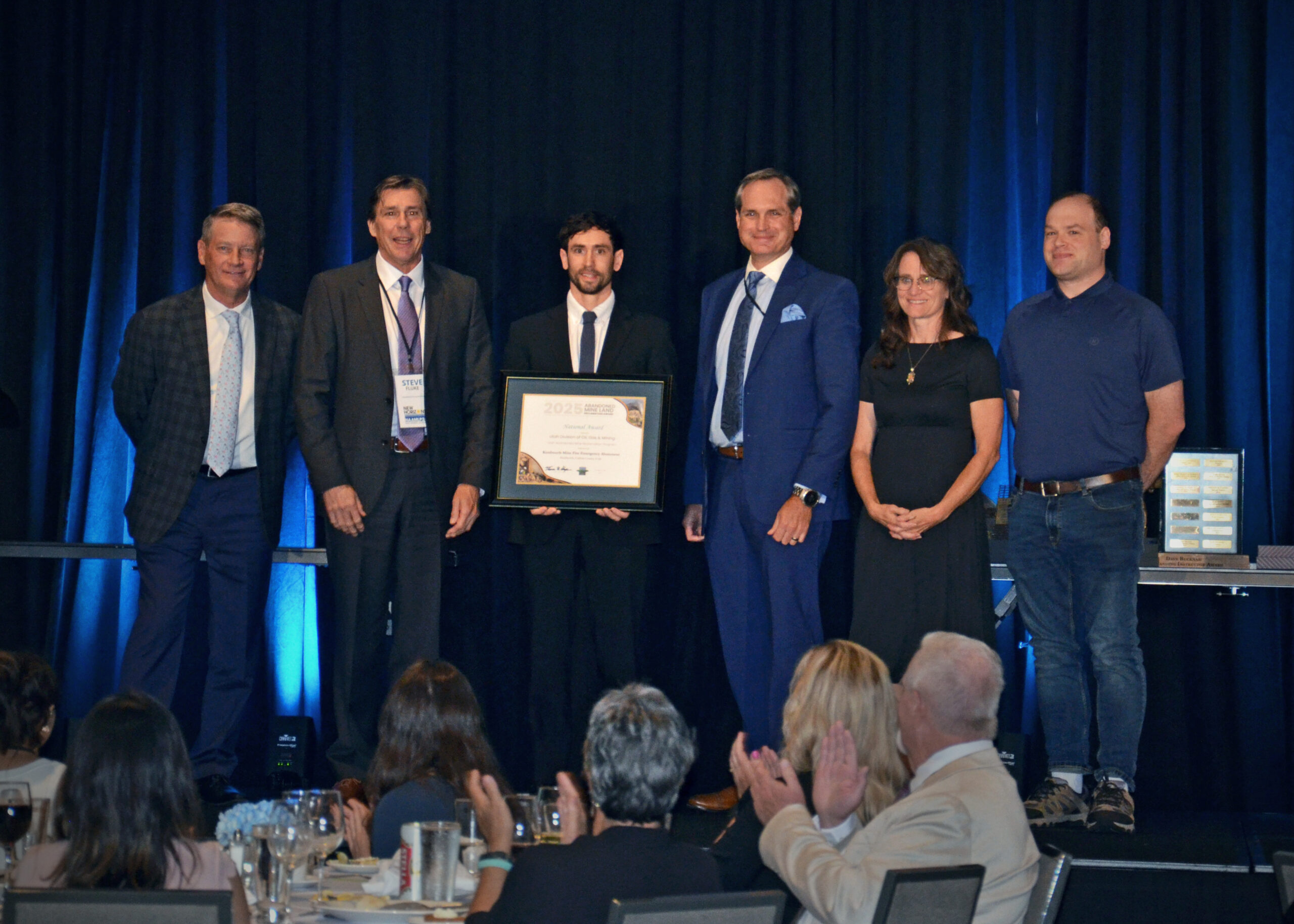 Close-up of a wooden boundary marker post in a rocky, disturbed earth landscape, typical of a mine site. The post is wrapped with bright yellow tape labeled 'BOUNDARY' and 'R647', indicating a new regulatory requirement.